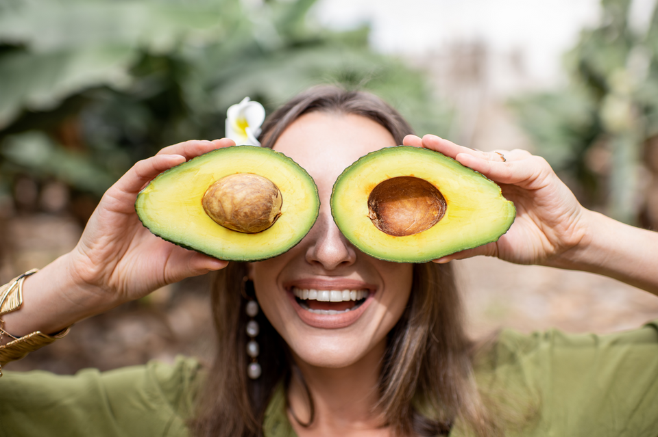 A healthy and smiling woman holds two halves of an avocado in front of her eyes. Green, natural, and blurred background. Concept of foods healthy for skin.