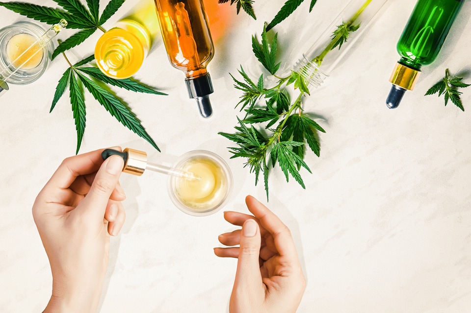 A pair of hands holding a dropper of CBD oil. Marijuana leaves and CBD bottles visible on the top edge. Concept of celebrities using CBD.