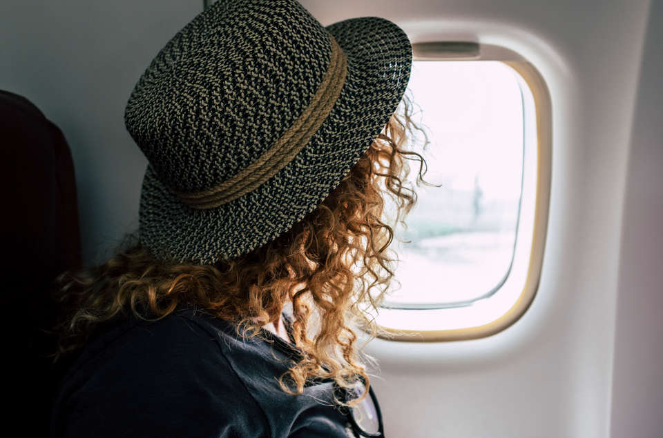 A woman with curly hair wearing a hat and looking out of a plane window, ready for her summer trip.